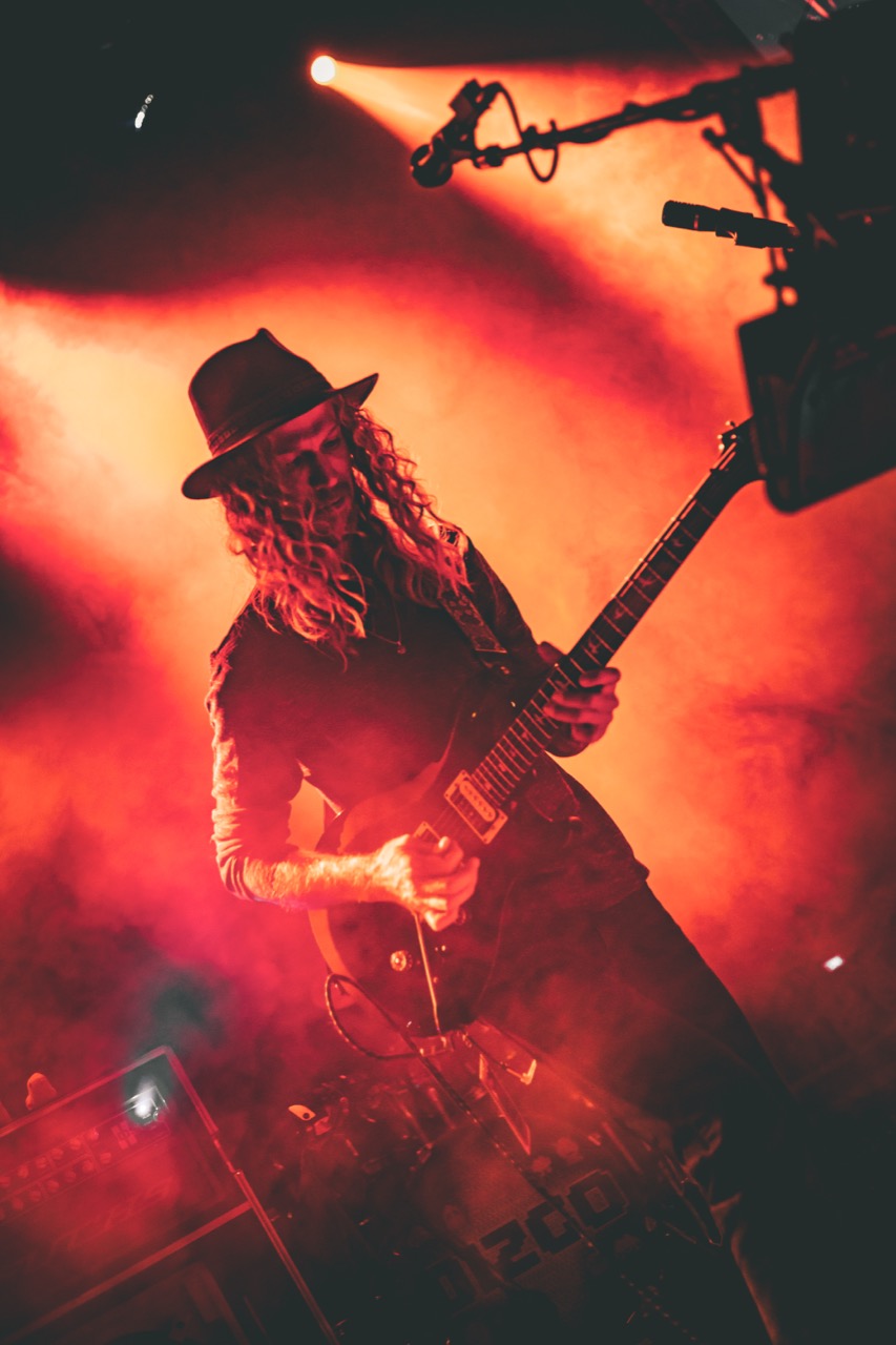 Dizgo guitarist in fedora and long hair playing under deep red smoky stage lighting