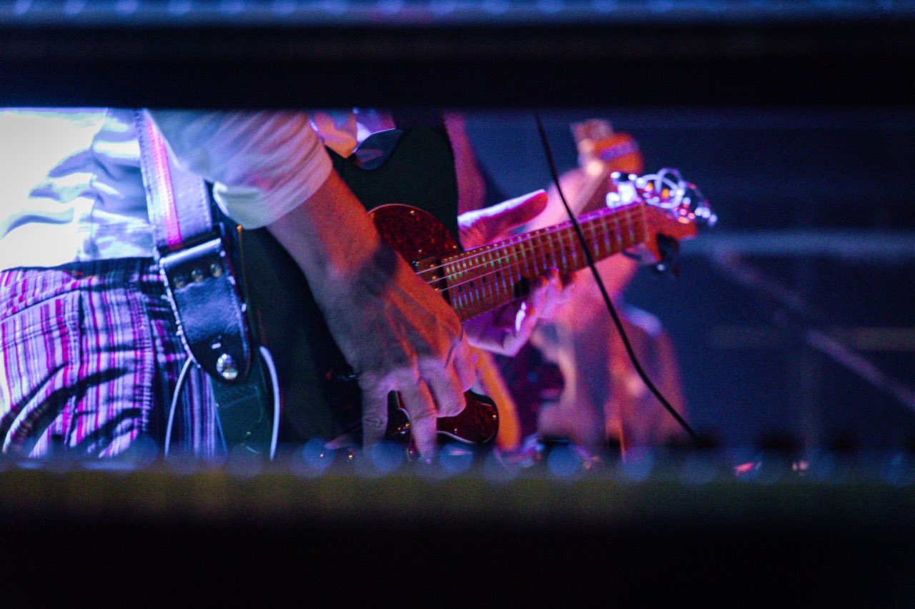 Close-up of a hand picking strings on a guitar with red tortoiseshell pickguard, shot through a stage frame