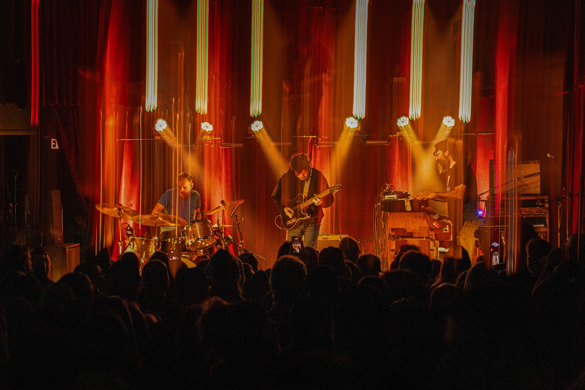 LaMP performing on stage with warm orange backlighting and vertical light strips, crowd silhouettes in foreground
