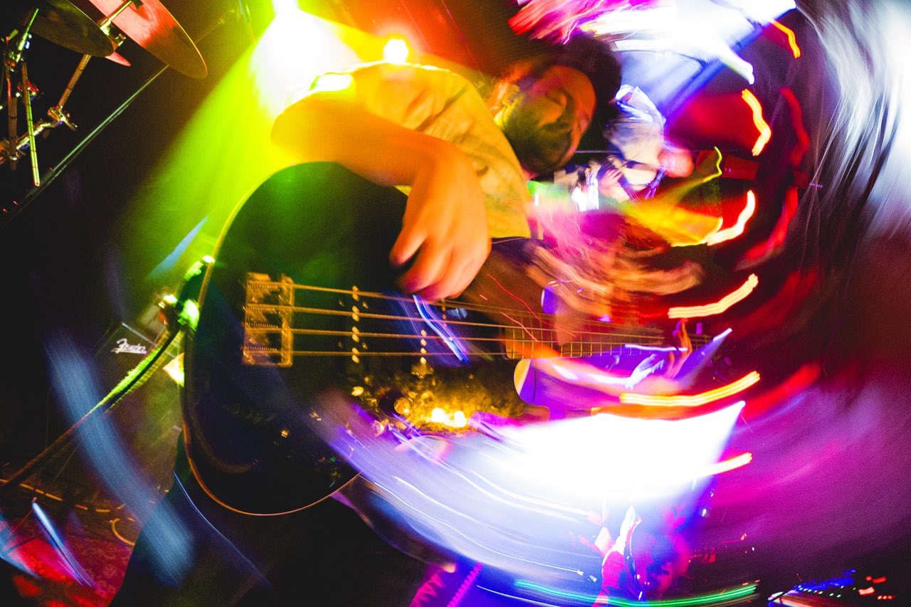 Mr. Please bassist captured with rainbow motion blur and long-exposure light streaks swirling around the bass body