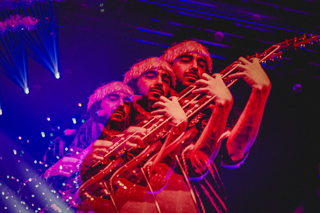 Mr. Please bassist captured in a triple-exposure under deep red stage lighting with blue laser highlights, three overlapping figures wearing fur hats