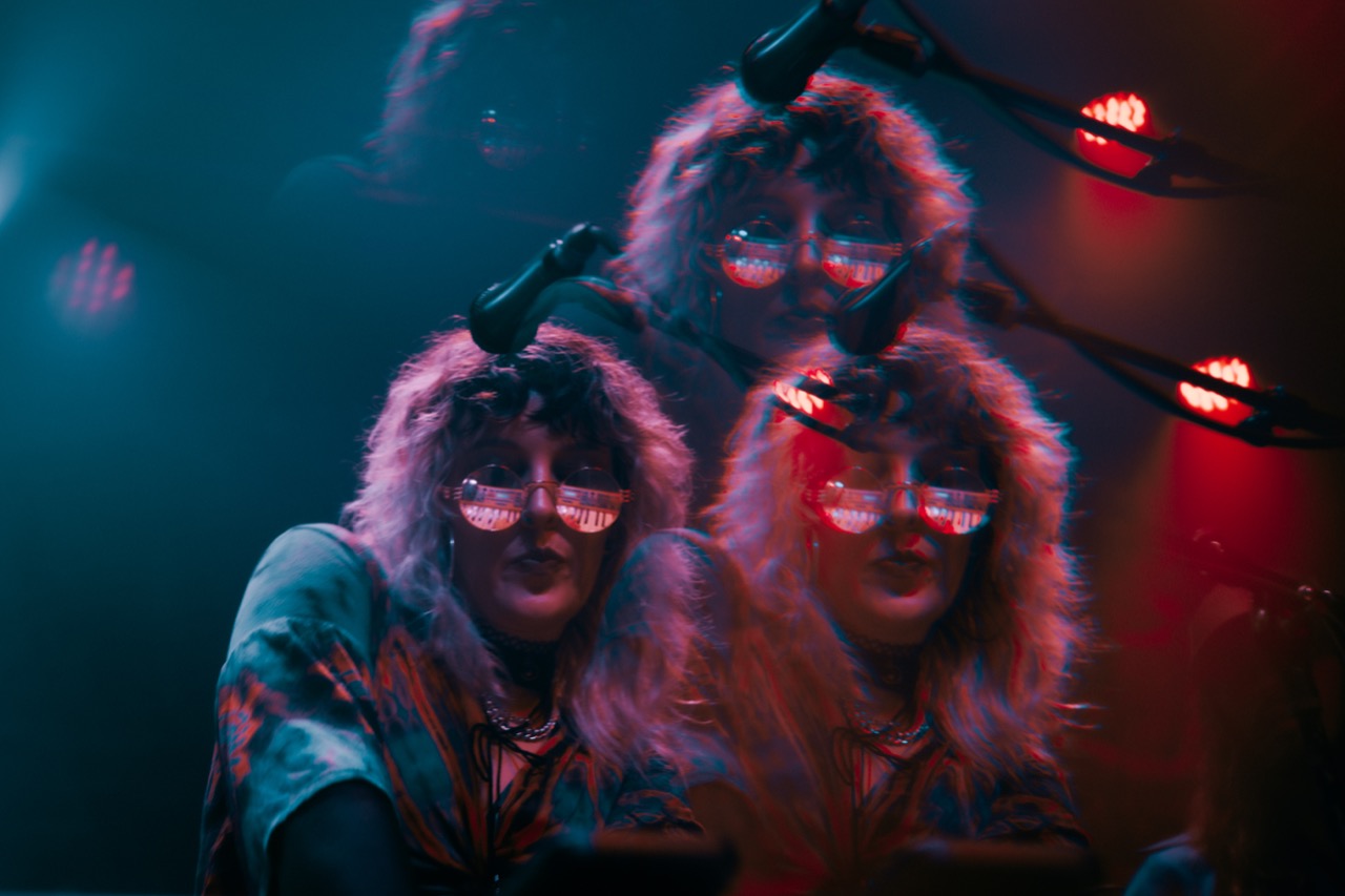 Natalie Brooke triple-exposure portrait wearing fur cap and pink-tinted glasses, red and teal lighting